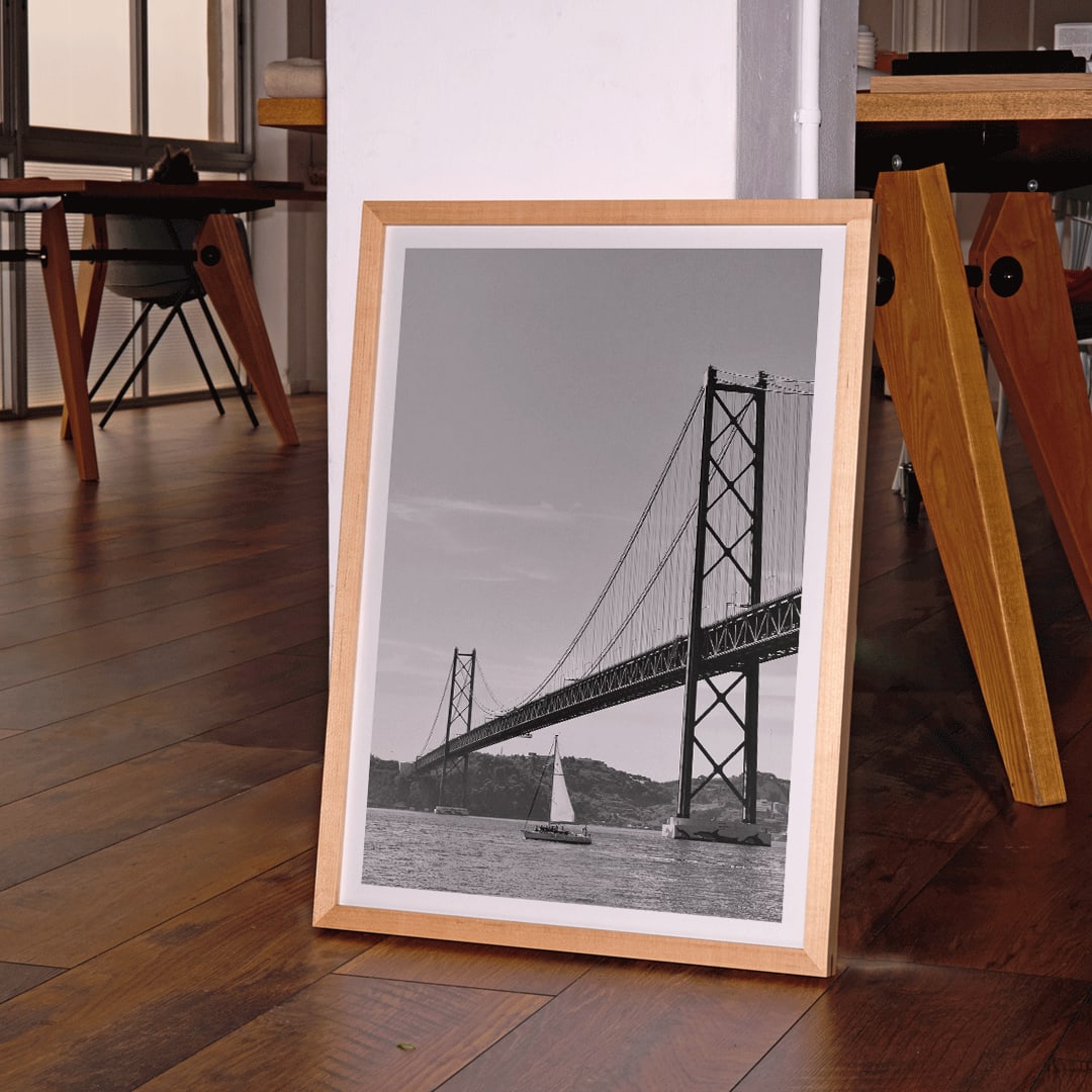 Lisbon Bridge Black and White Poster by Raphael Alves featuring a framed photograph of a suspension bridge and sailboat in the water, framed and displayed in home decor