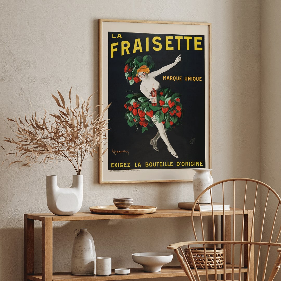 The Fraisette by Leonetto Cappiello poster featuring a whimsical woman in a strawberry costume holding a bottle of liqueur against a black background, framed and displayed in home decor